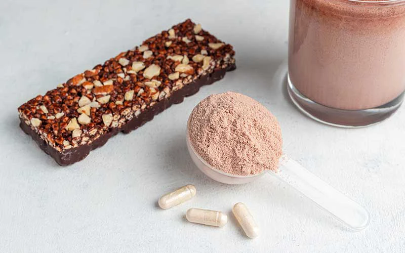 Scoop of whey protein isolate, chocolate milkshake in a glass, granola bar, white capsules