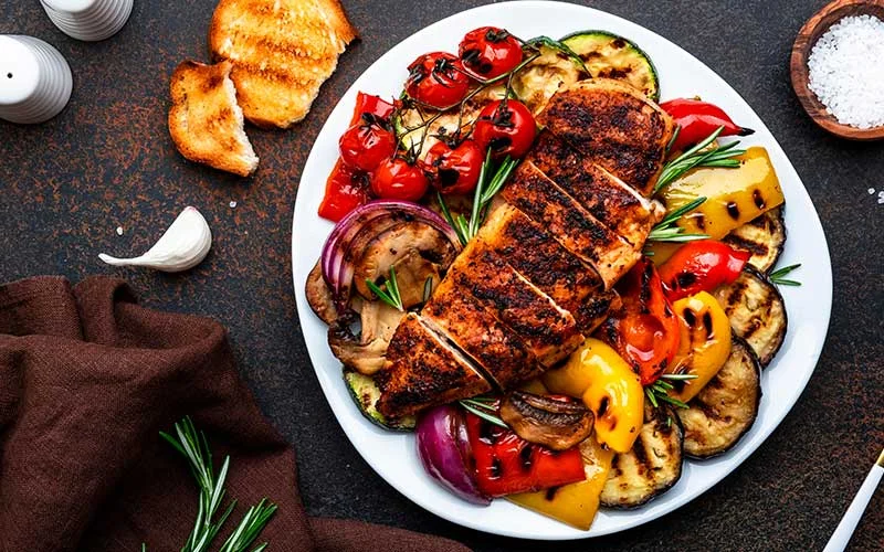 Grilled chicken filet and vegetables salad. Colorful paprika, zucchini, eggplant, mushrooms, tomatoes, red onion with rosemary, served on plate, brown table background
