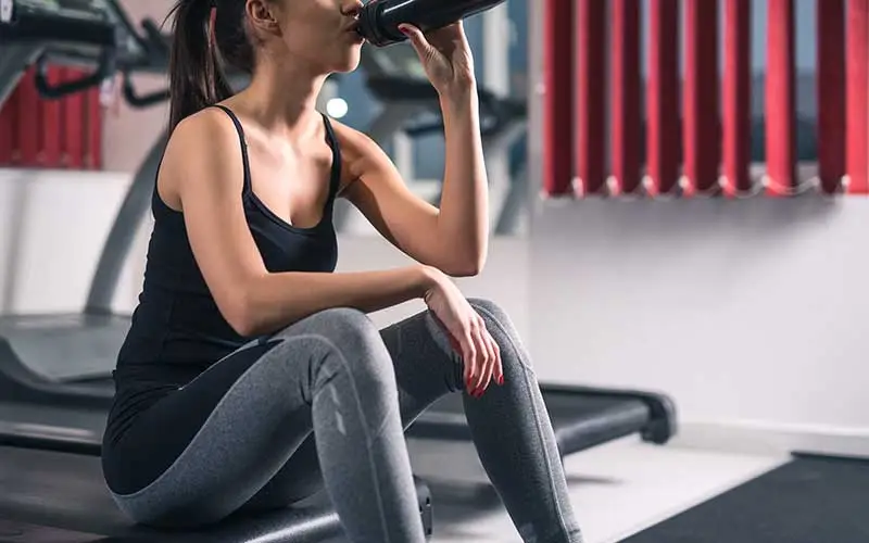 Sporty young woman drinking meal replacement shake from a bottle while sitting on a treadmill at the gym