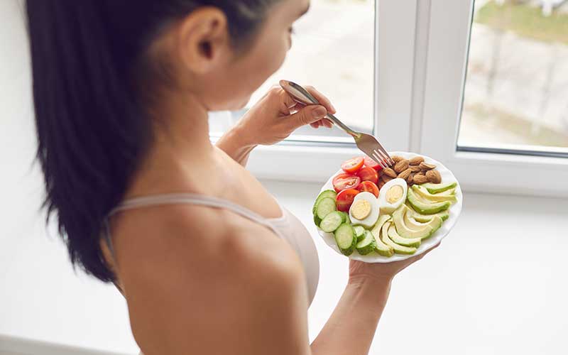 woman athlete in sportswear standing and eating a healthy meal with eggs, nuts and vegetables after a workout at home