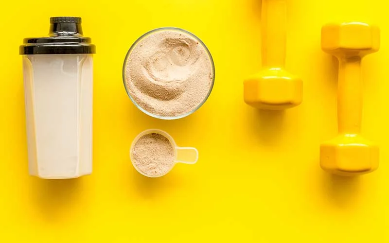 Meal replacement shake, scoop and dumbbells on yellow background