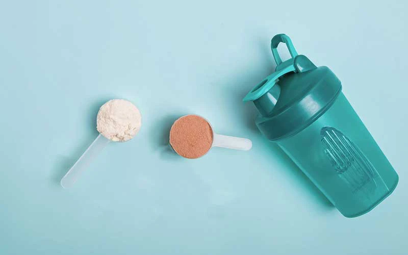 Scoops of chocolate and vanilla meal replacement shake and shaker on cyan blue background.