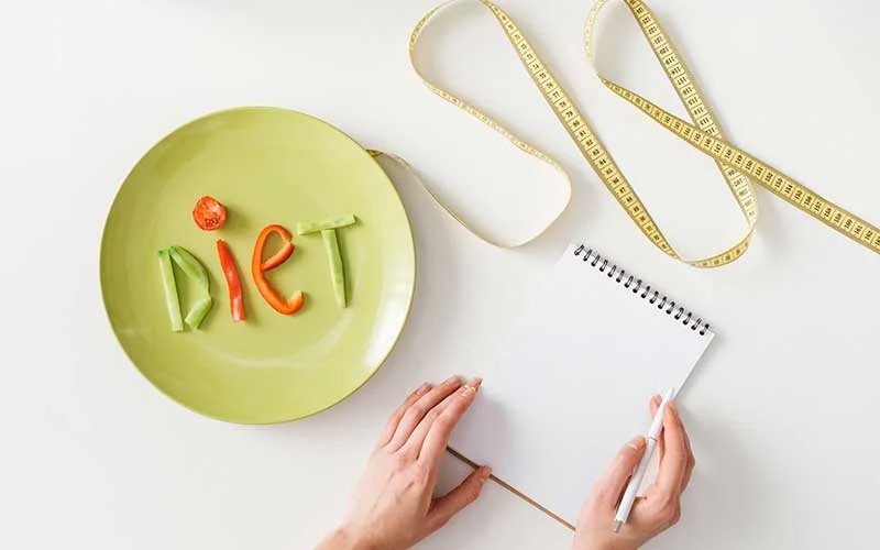 Woman writing in notebook near measuring tape and plate with diet lettering from vegetable slices on white background