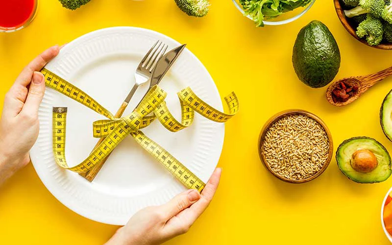 Fork and knife tied with measure tape on plate in hands on yellow background with vegetables.