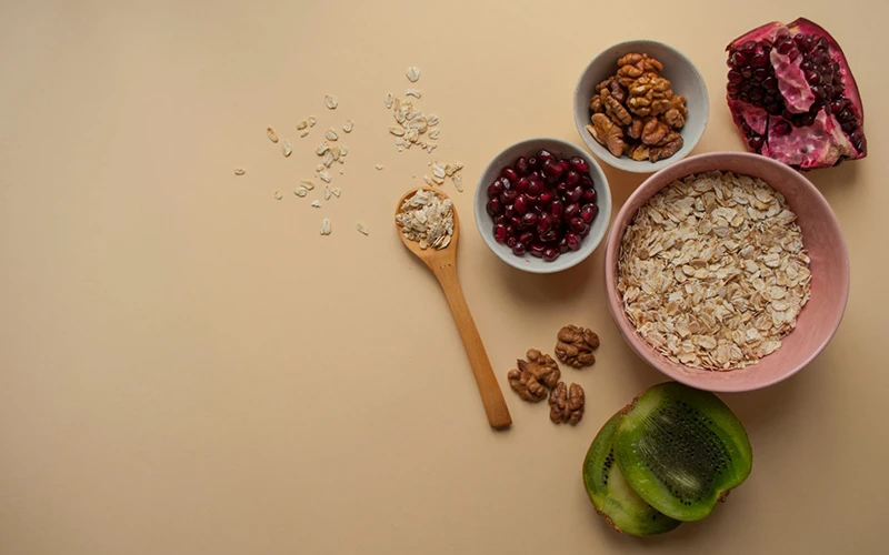 Oatmeal, pomegranate seeds, kiwi fruit, and superfoods as healthy breakfast ingredients on beige colored background.