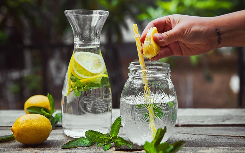 Hand squeezing lemon into a glass jar with water side view on wooden and yard table.
