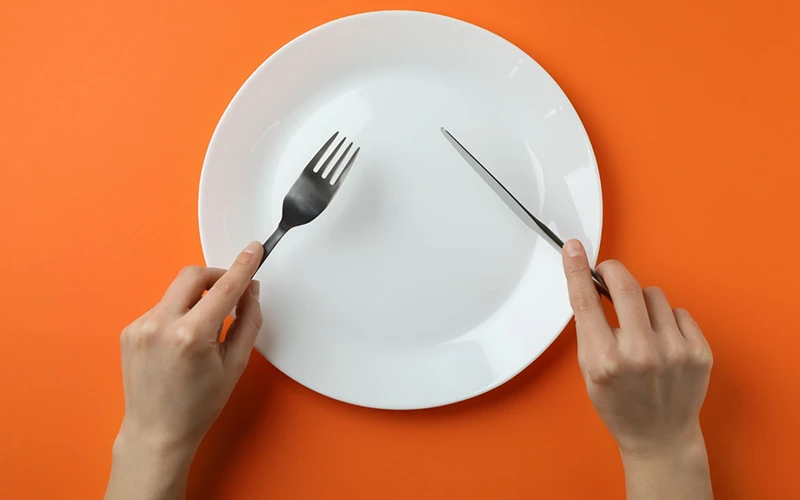 Woman holding fork and knife near empty plate on orange background