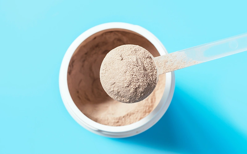 Chocolate meal replacement shake powder in a scoop against a plastic jar on blue background